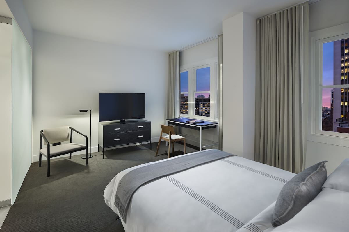 A hotel room with a bed, a desk, a chair, a television, and a window with a city view.