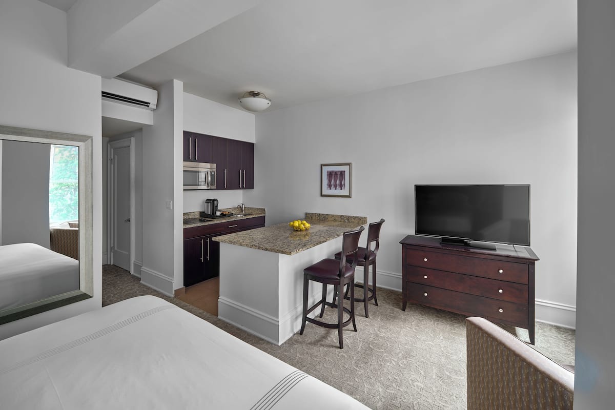 A modern hotel room with a separate kitchen area, a dining space, a TV, and a large bed.