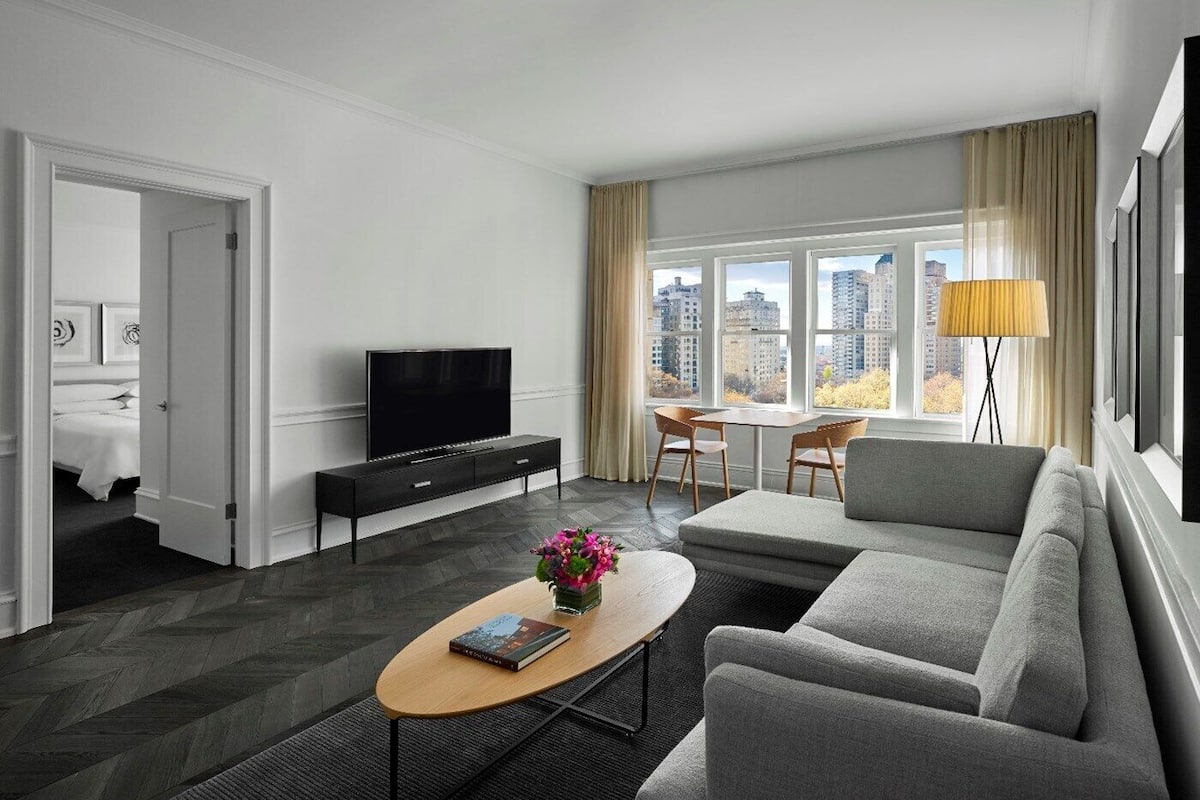 A modern hotel room with a large window, a flat-screen TV, a sofa, a coffee table with a book, and a dining area with chairs.