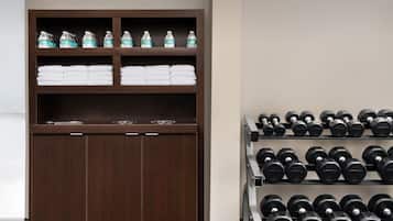 A wooden cabinet with water bottles and folded towels on the top shelf, and a rack of black dumbbells below.