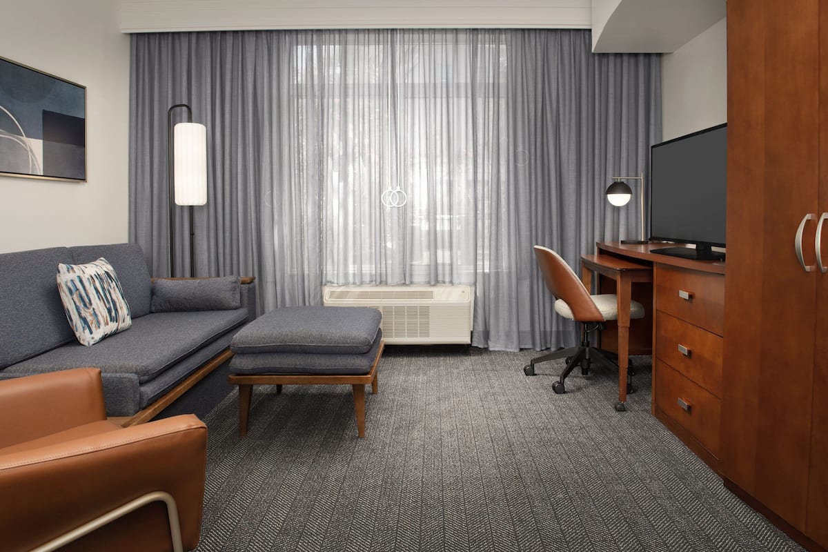 A modern hotel room with a grey sofa, a wooden desk, a television, and a large window with grey curtains.