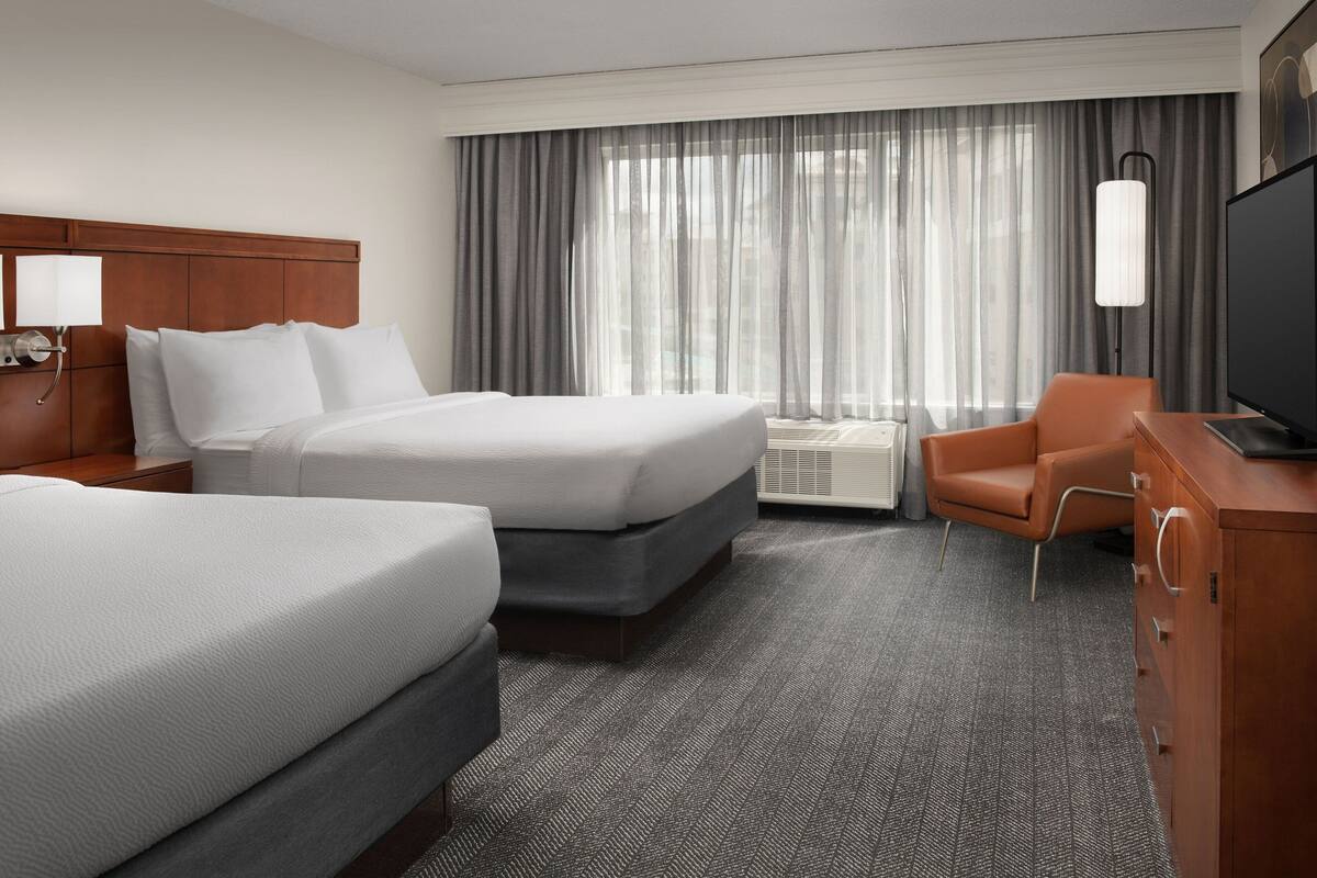 A hotel room with two beds, a desk, a chair, a TV, and a large window with curtains.