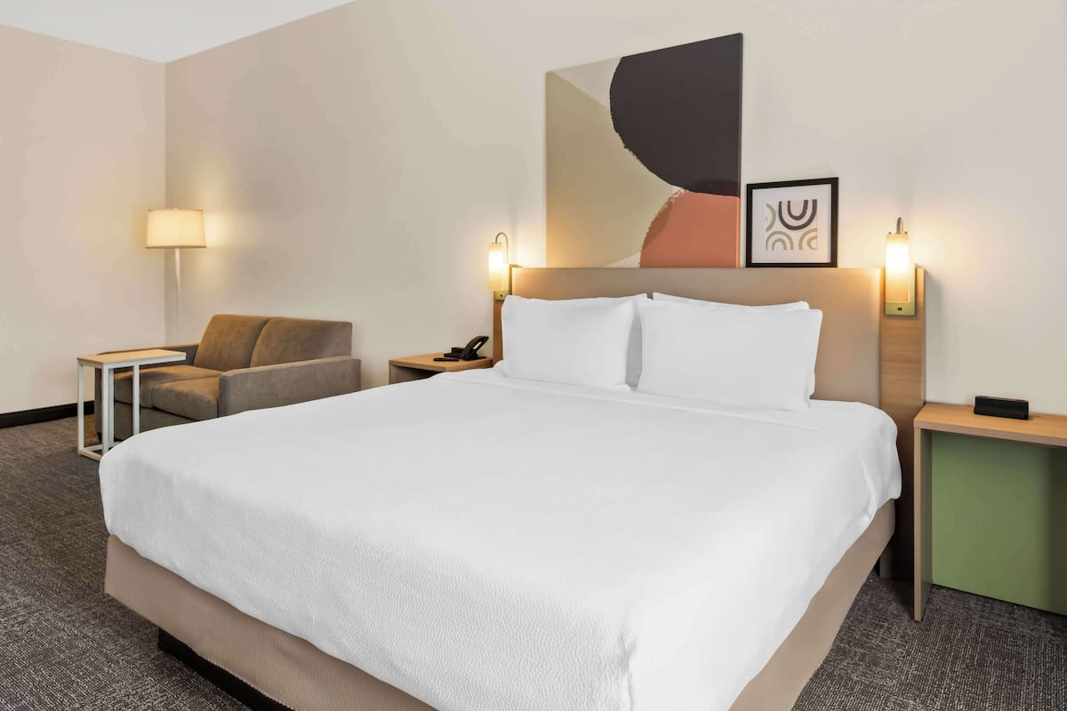 A hotel room with a large bed, a chair, a nightstand, a lamp, and a wall-mounted artwork.