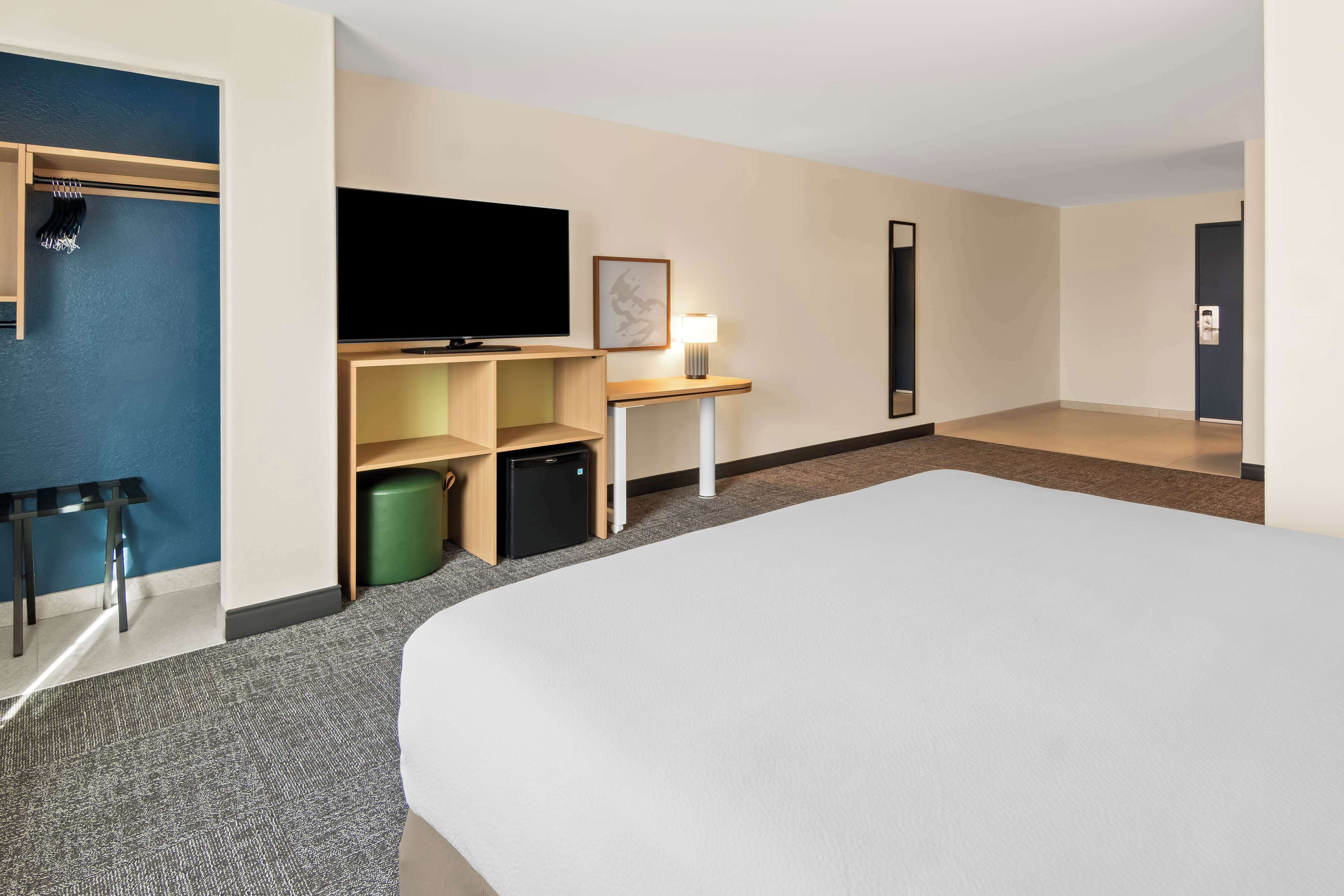 Ein Hotelzimmer mit einem großen Bett, einem Flachbildfernseher, einer kleinen Kühlbox, einem Schreibtisch mit Lampe, einem Kleiderständer und einem Mülleimer.
