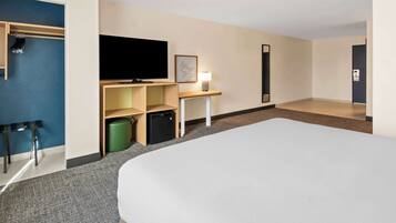 Ein Hotelzimmer mit einem großen Bett, einem Flachbildfernseher, einer kleinen Kühlbox, einem Schreibtisch mit Lampe, einem Kleiderständer und einem Mülleimer.