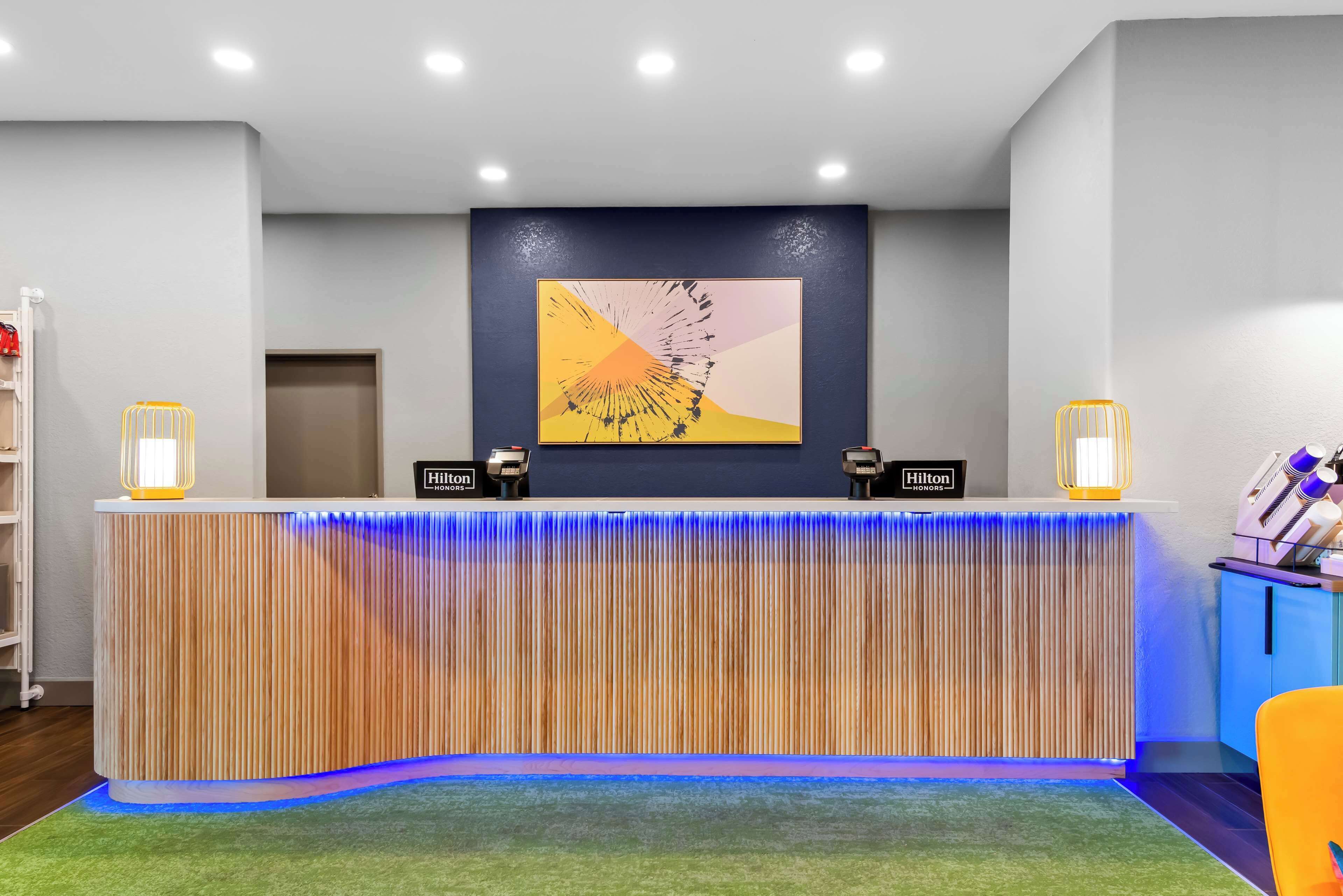 A hotel reception desk with a modern design, featuring a vertical wooden panel, a colorful abstract painting above, and two illuminated wall sconces.