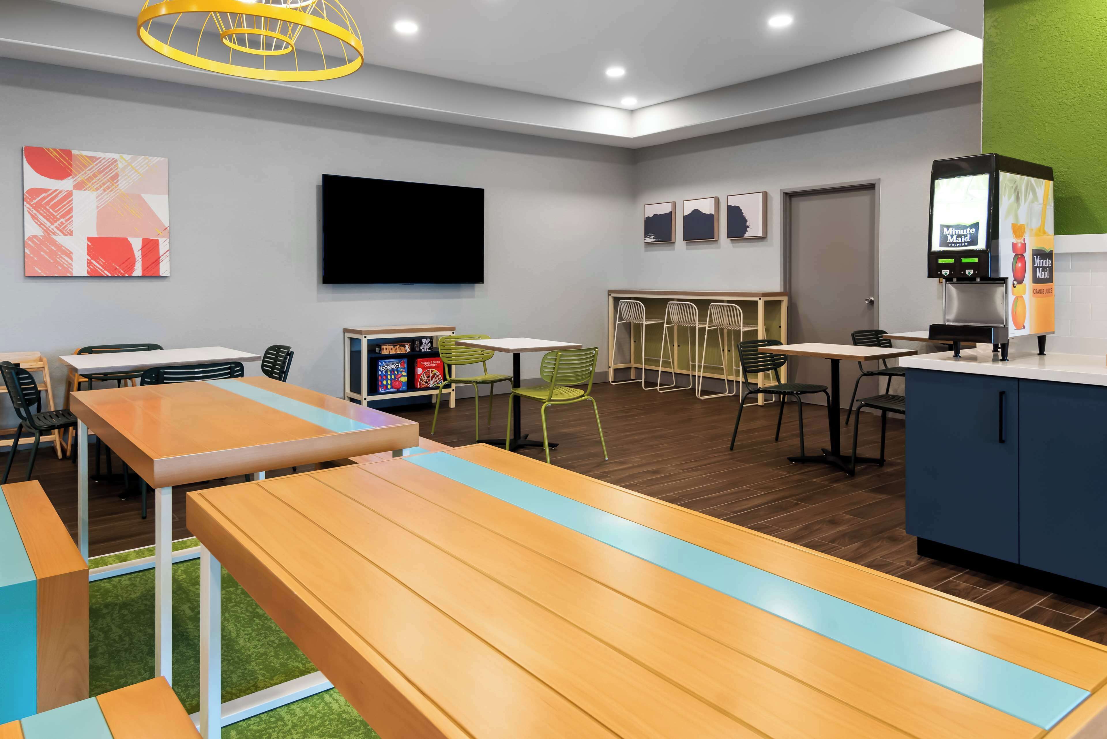 Un espace repas moderne avec des tables et des chaises en bois, une machine à café et un téléviseur à écran plat fixé au mur.