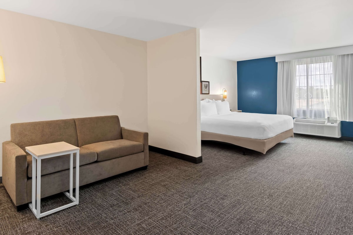 A hotel room with a beige sofa, a small white side table, a bed with white linens, a nightstand, a wall-mounted lamp, and a window with sheer curtains.