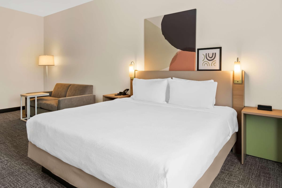 A hotel room with a large bed, a chair, a desk, and a wall-mounted artwork.