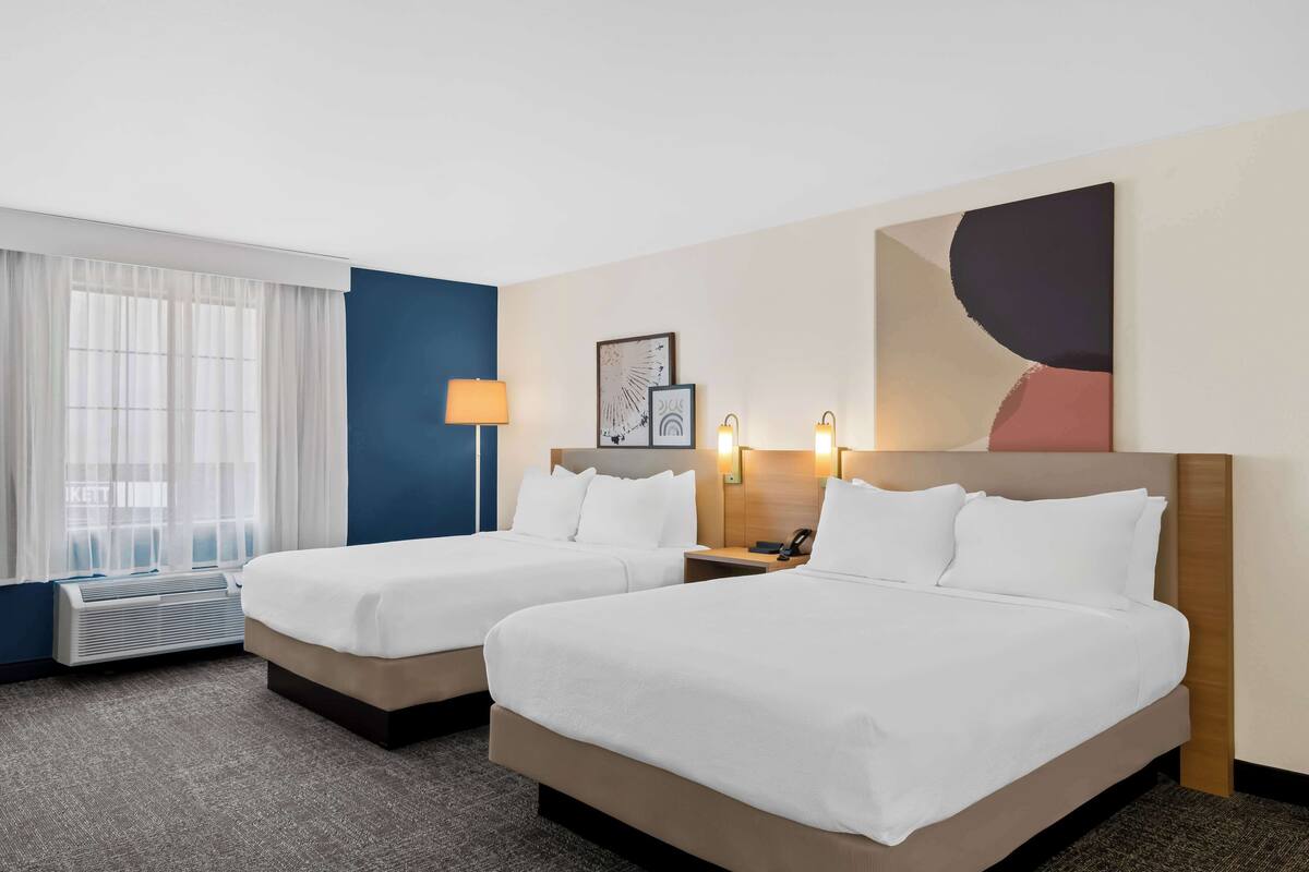 A hotel room with two beds, a large window with curtains, a wall-mounted artwork, and a bedside table with a lamp.