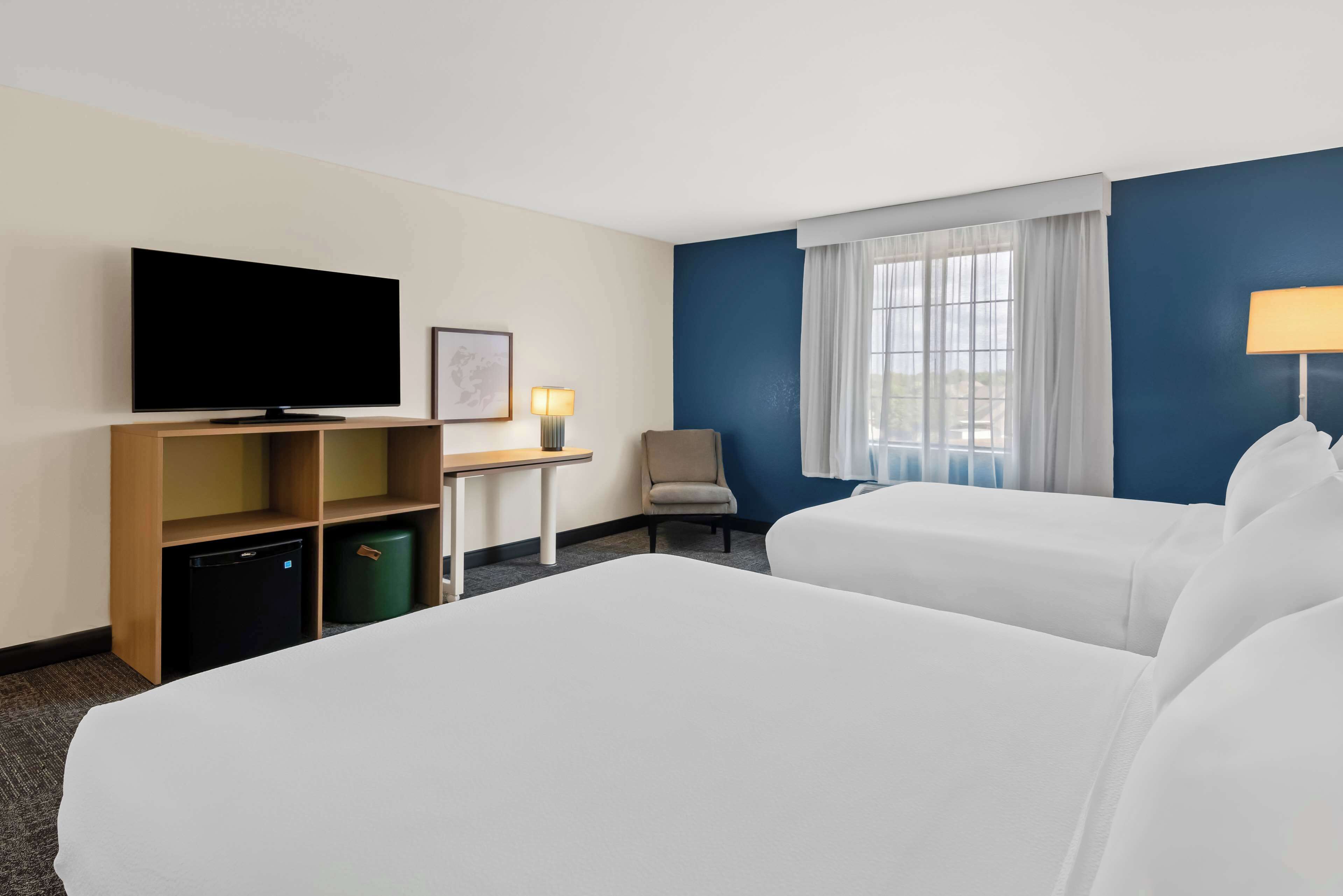 A hotel room with two beds, a television, a desk with a lamp, a chair, and a window with curtains.