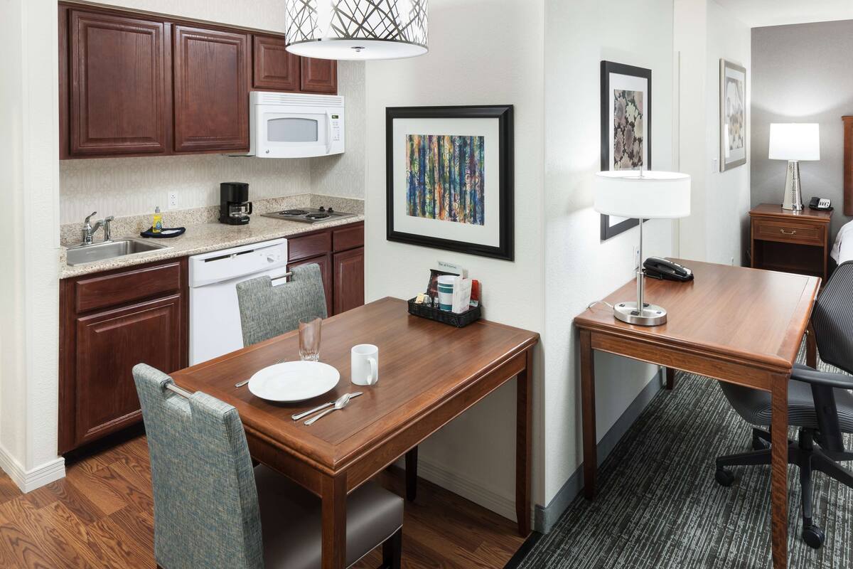 A compact hotel room with a kitchenette, a dining area with a wooden table and chairs, a small desk with a lamp, and a bed with a headboard.