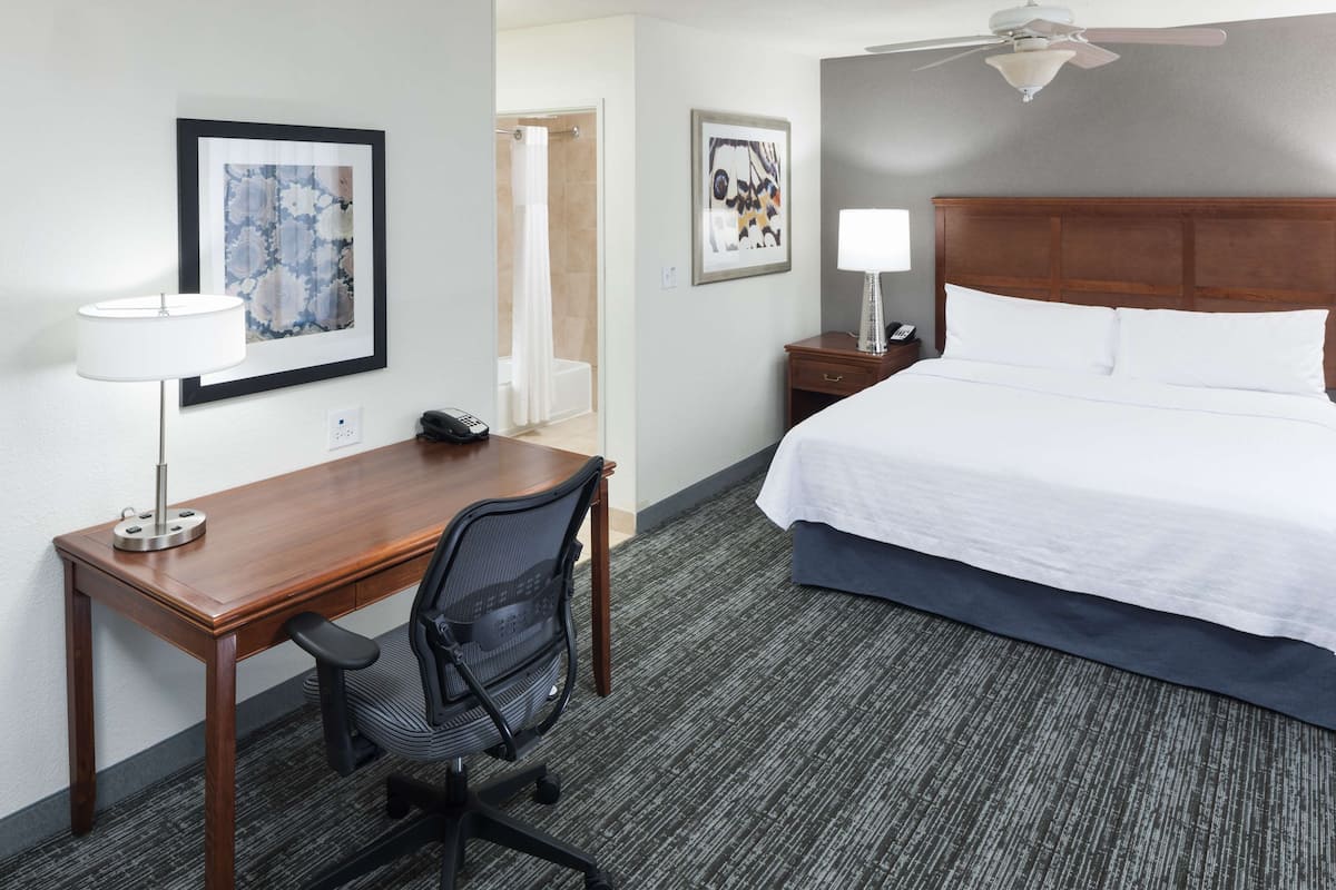 A hotel room with a wooden desk, a black office chair, a bed with white linens, a ceiling fan, and two framed artworks on the walls.