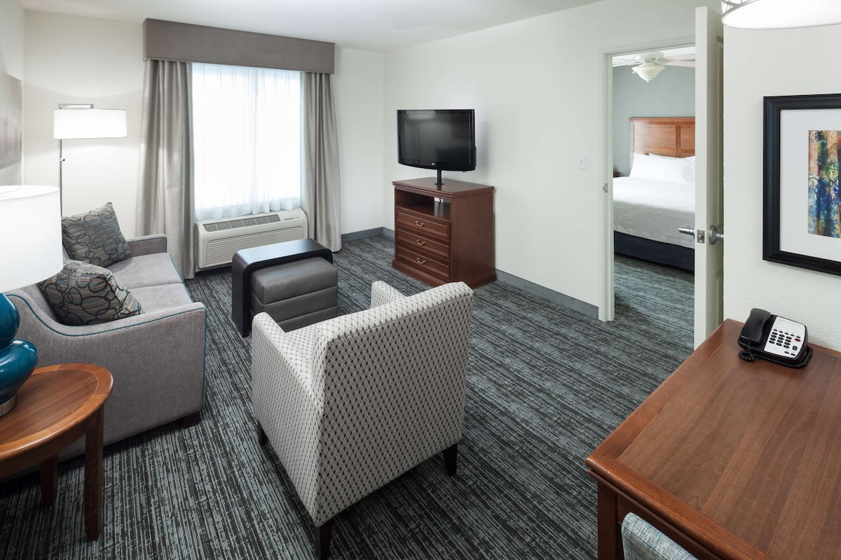 A hotel room with a sofa, armchair, desk, TV, and a bed in the adjacent room.
