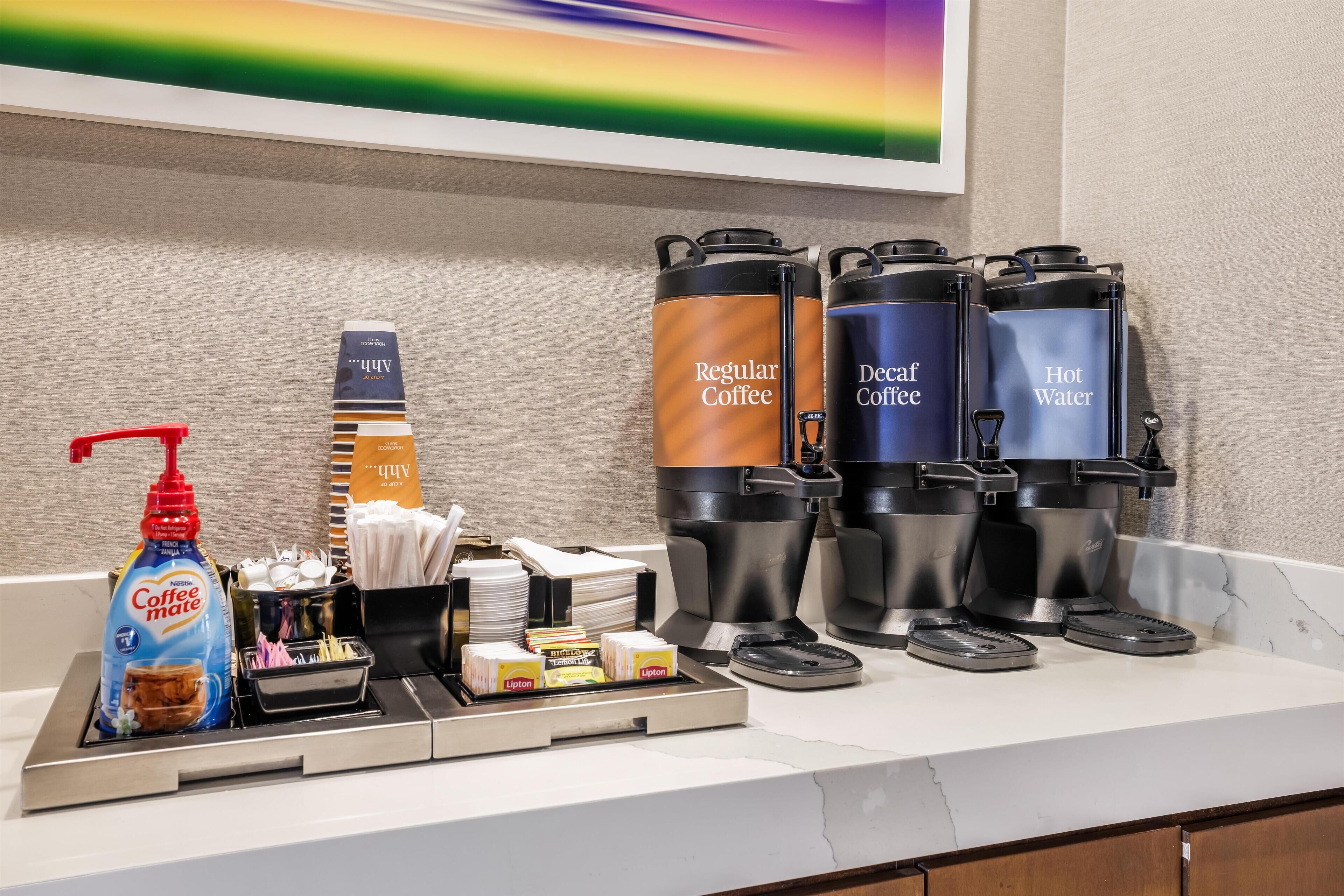A coffee station with dispensers labeled 'Regular Coffee,' 'Decaf Coffee,' and 'Hot Water,' along with a bottle of Lipton tea and other amenities.