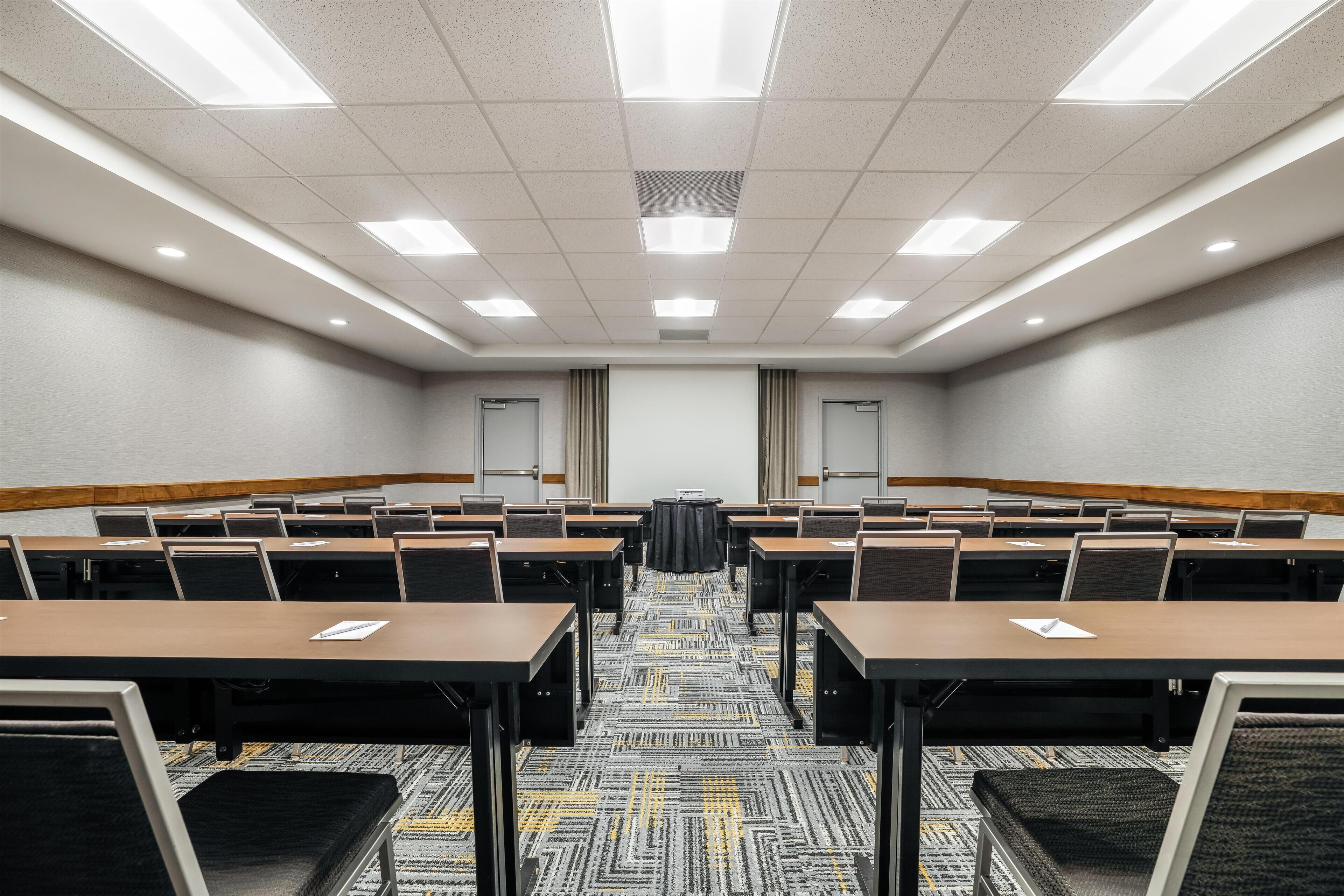 A conference room with multiple tables and chairs, a projector screen, and a carpeted floor.
