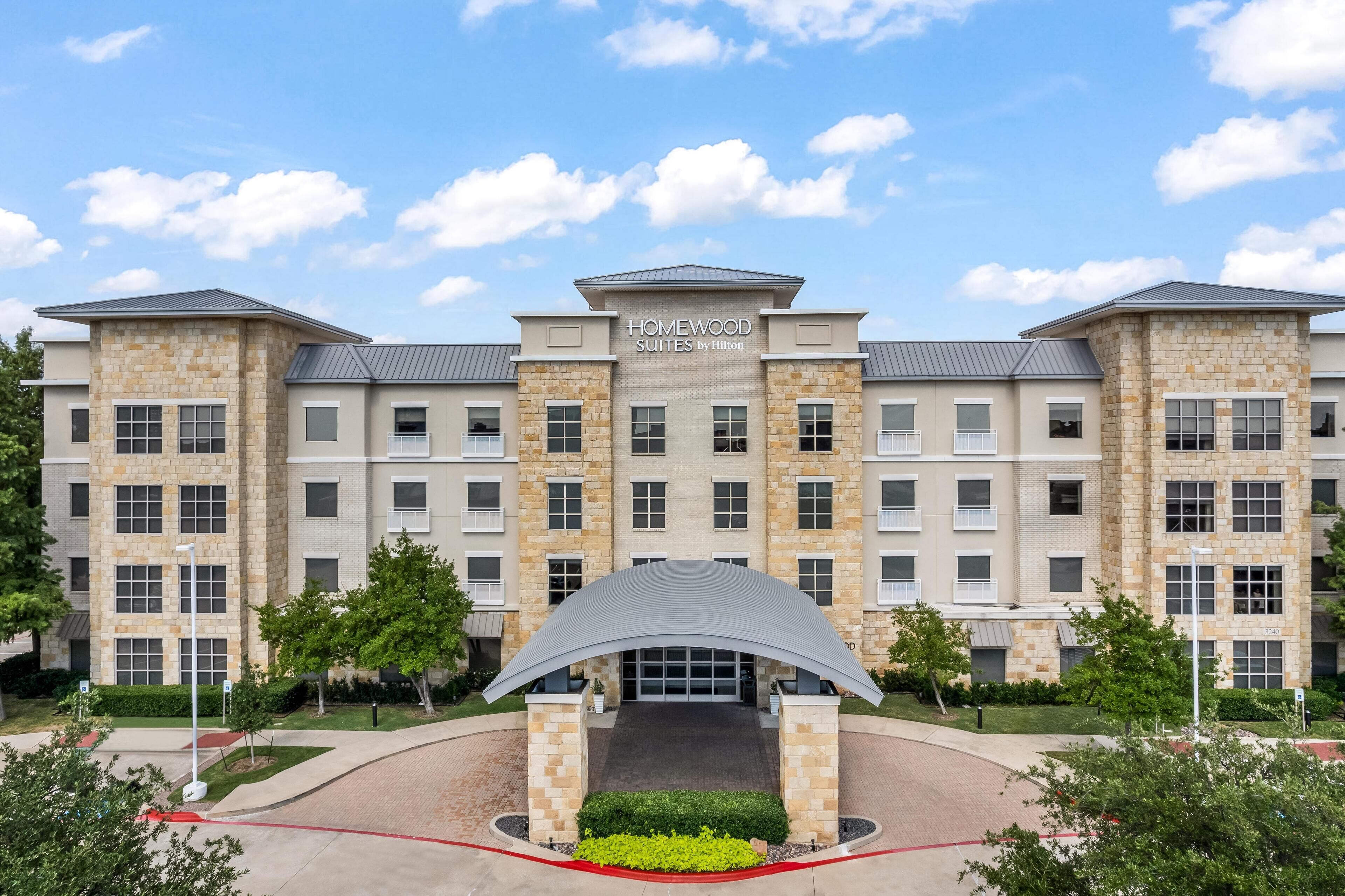 A multi-story hotel with a stone facade, a central entrance with a curved roof, and a clear sky.