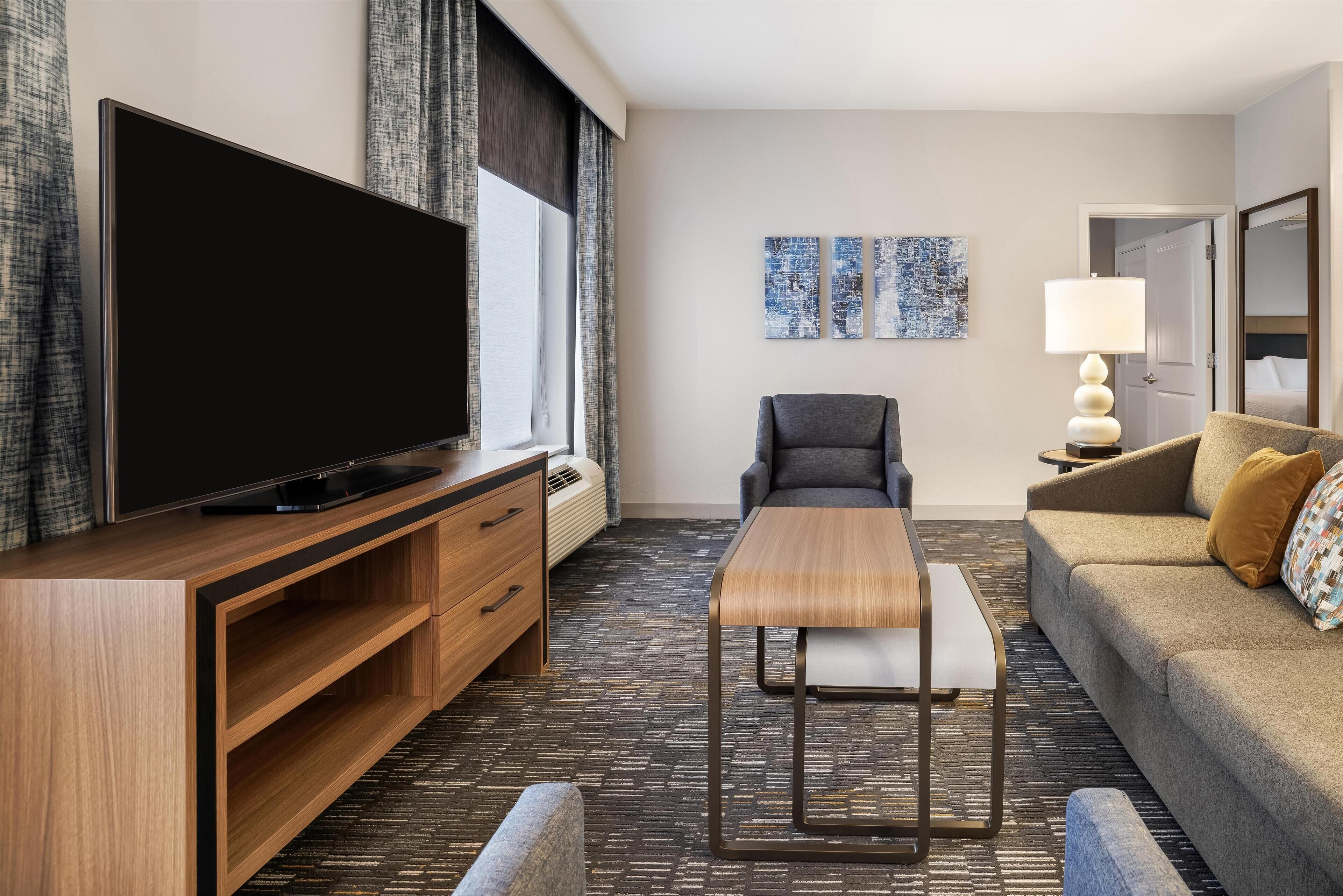 Habitación de hotel moderna con una televisión de pantalla plana, un mueble de entretenimiento de madera, un sofá gris, una mesita pequeña y una lámpara.