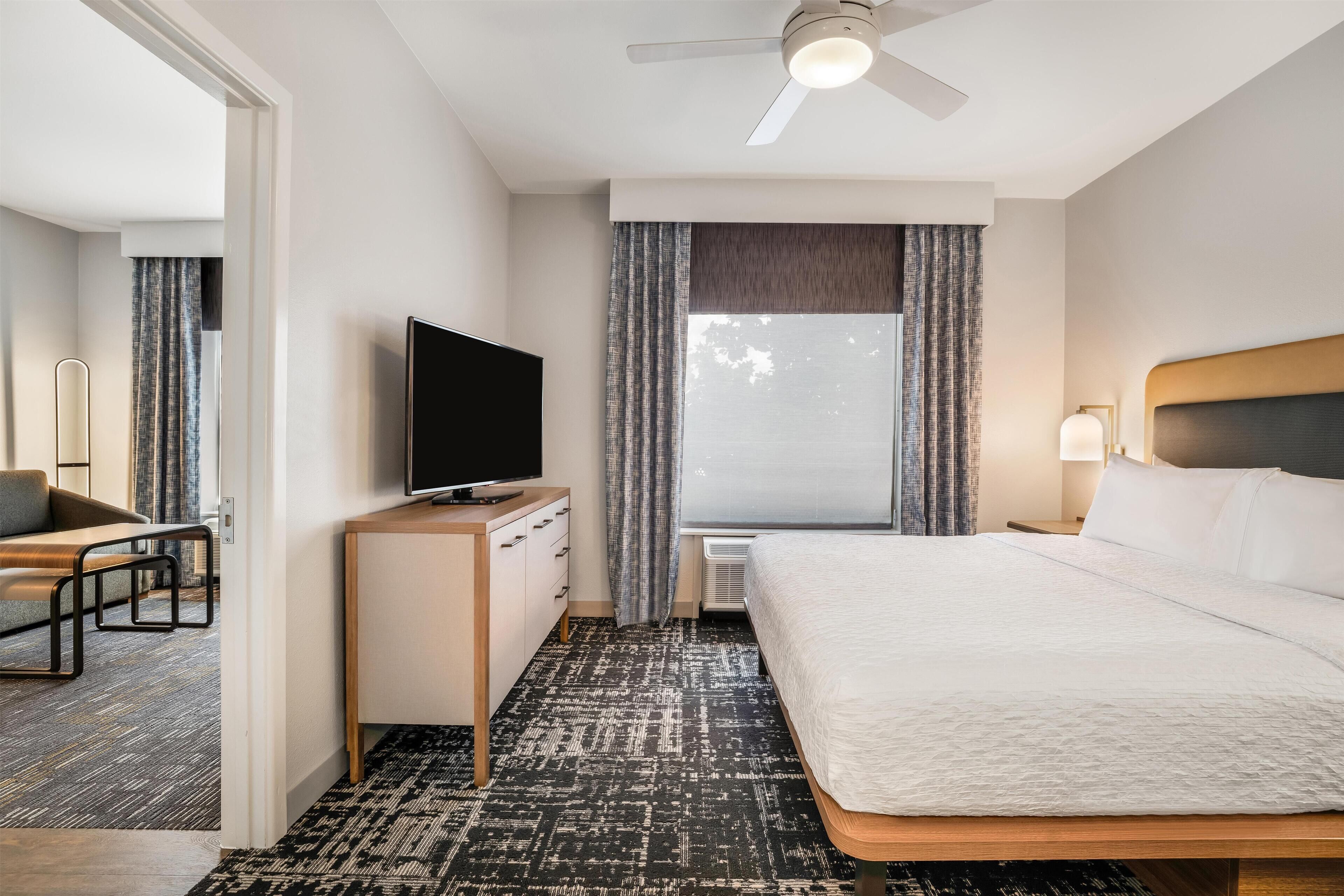 Une chambre d’hôtel avec un lit, un bureau avec une télévision, un ventilateur de plafond et une fenêtre avec des rideaux.