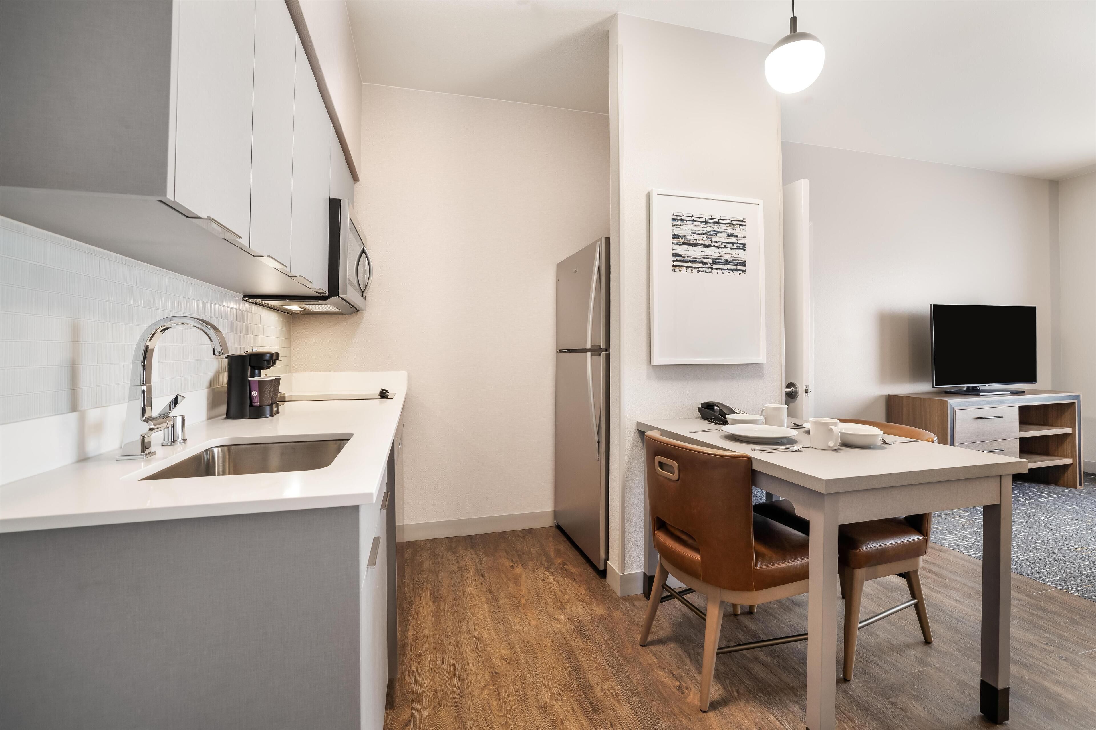 A modern kitchen with a built-in refrigerator, a coffee machine, and a sink. There is a dining area with a table and chairs. A flat-screen TV is visible in the background.