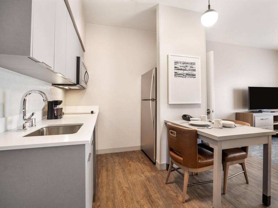 A modern kitchen with a built-in refrigerator, a coffee machine, and a sink. There is a dining area with a table and chairs. A flat-screen TV is visible in the background.