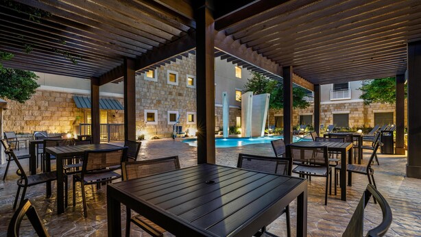 An outdoor patio area with wooden tables and chairs, a stone wall, and a pool in the background. - Homewood Suites by Hilton Dallas-Frisco (Frisco)