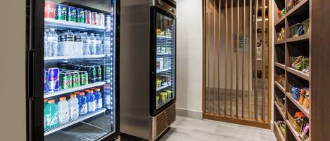 Un pasillo con dos dispensadores refrigerados de bebidas, repletos de diversas opciones, una puerta de madera y un estante con aperitivos.
