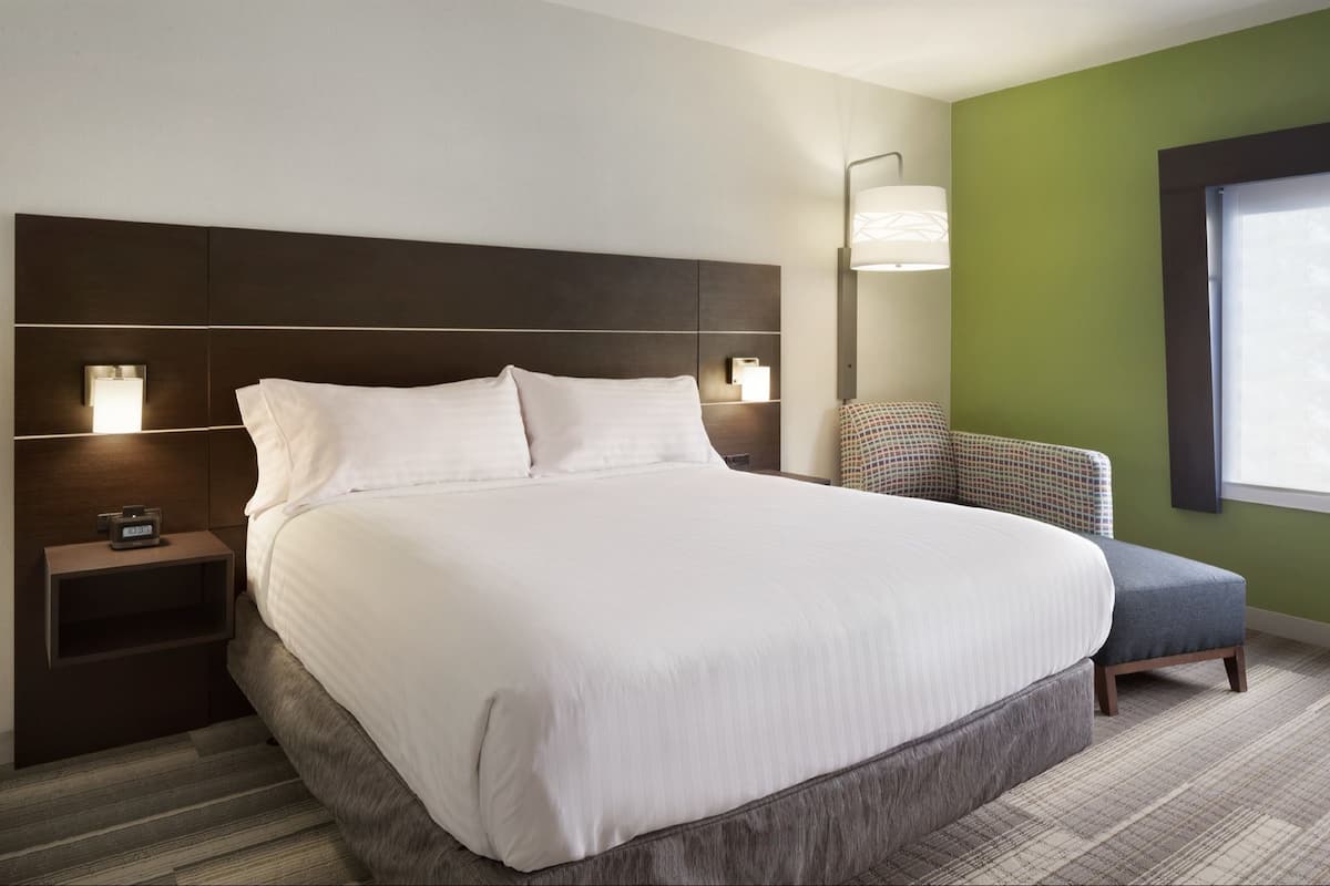 A large bed with a dark headboard, bedside lamps, a small table with a clock, and a chair with a patterned cushion.