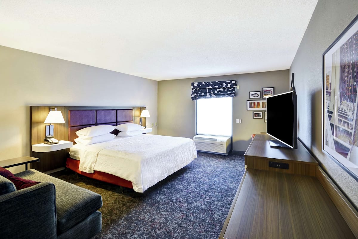 A hotel room with a bed, bedside tables, a TV, a chair, a window with a view, and a wall-mounted TV.