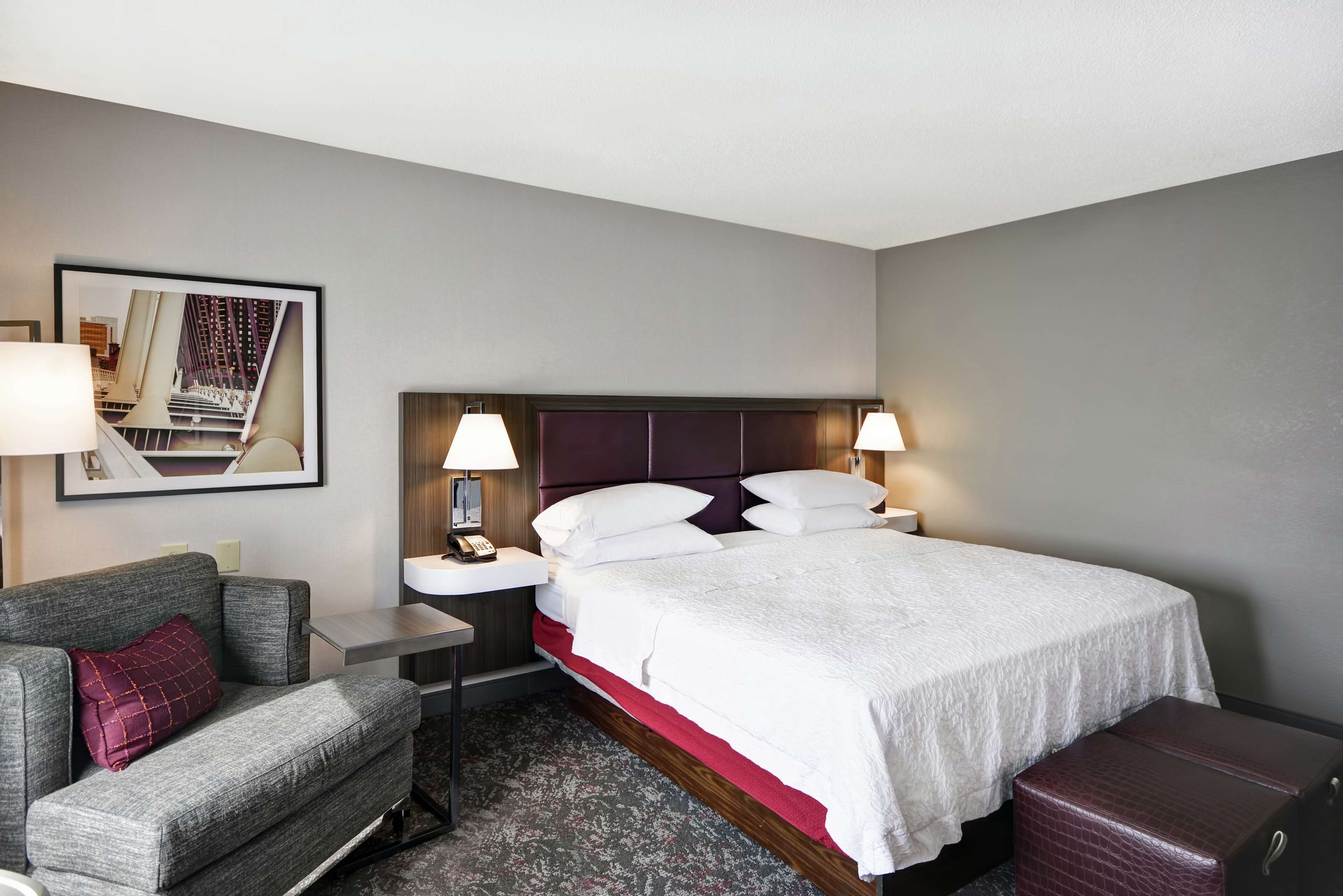 A hotel room with a bed, a nightstand, a lamp, a chair, and a suitcase.