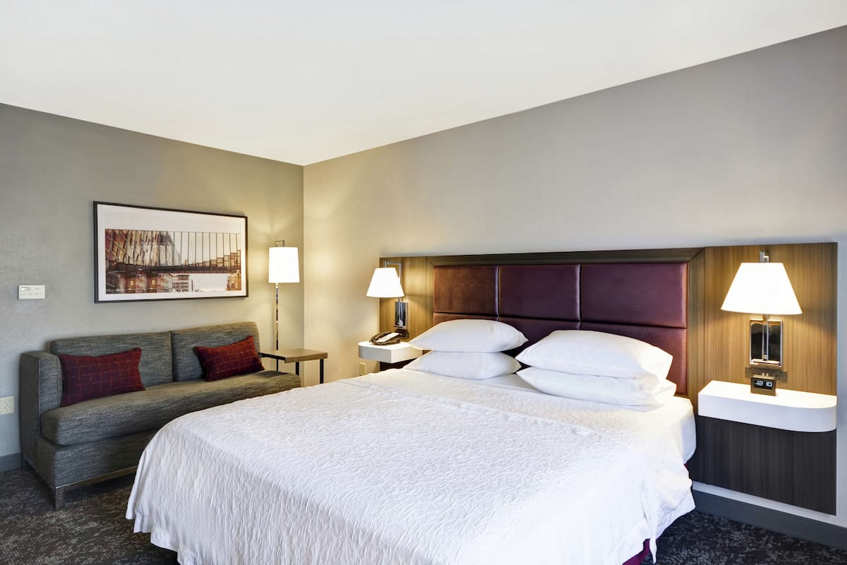 A hotel room with a large bed, two bedside tables with lamps, a sofa with red cushions, a framed picture on the wall, and a wooden headboard.