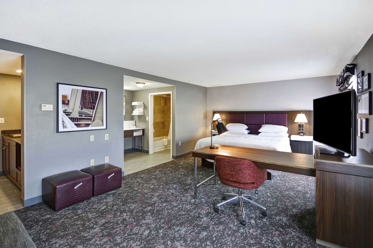 A hotel room with a bed, desk, chair, and a television. There is a framed picture on the wall and a small seating area with two ottomans.