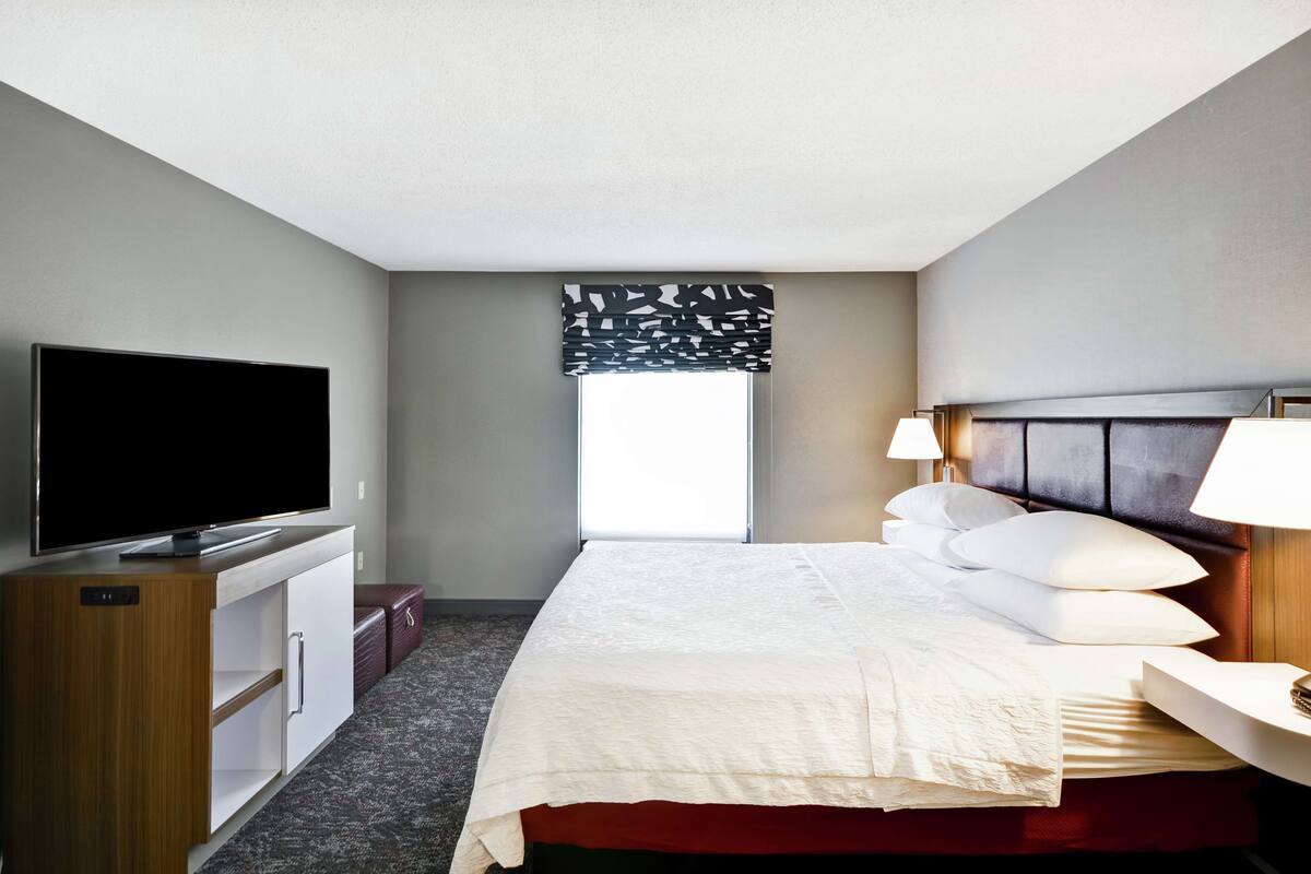 A hotel room with a bed, a TV, a desk with a lamp, a window with blinds, and a carpeted floor.