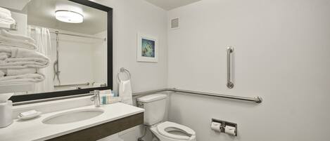 A modern bathroom with a white sink, mirror, and toilet. There are handrails on the walls and a towel rack with folded towels.