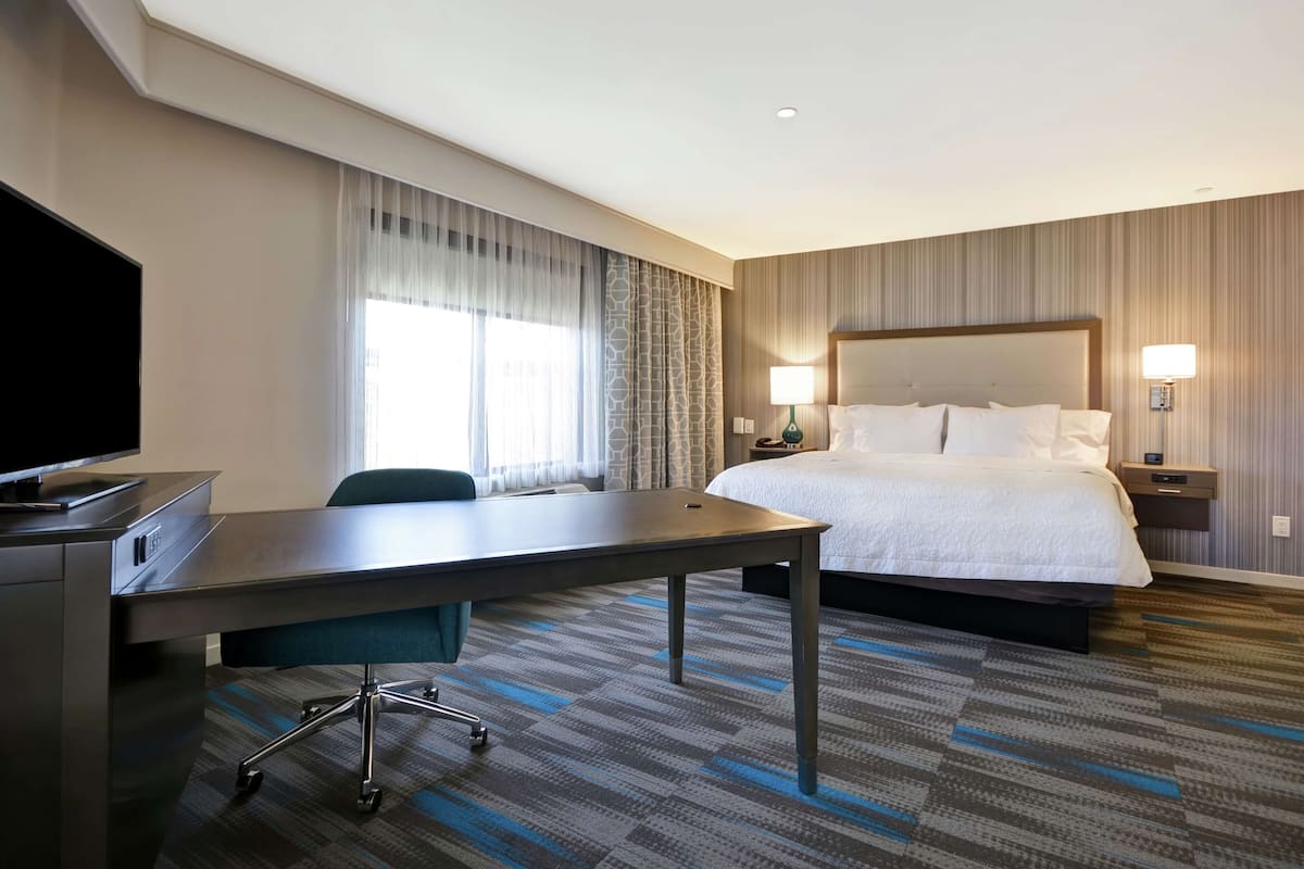 A hotel room with a bed, a desk with a chair, a TV, and a window with curtains.