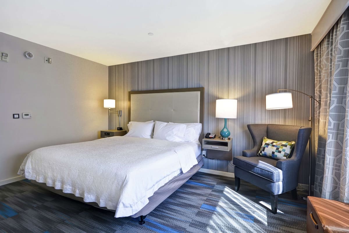 A hotel room with a large bed, two bedside tables with lamps, a chair, and a wooden nightstand.