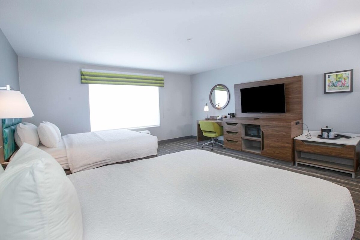 Habitación de hotel con una cama grande, un escritorio, una silla, un televisor y una ventana con cortina verde.