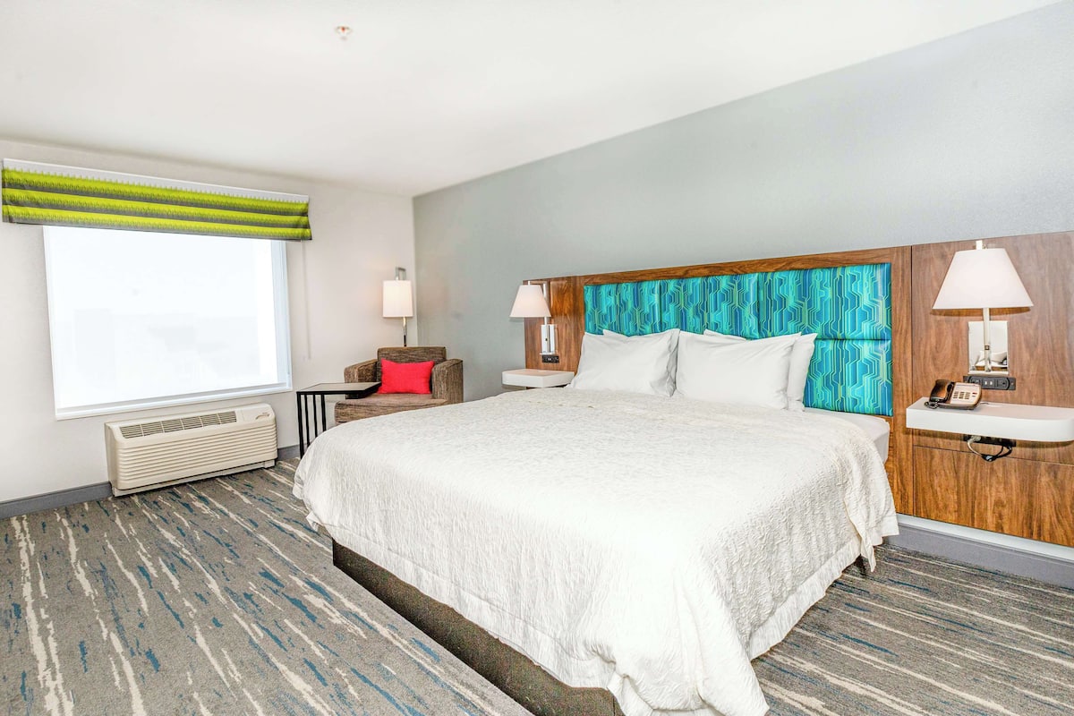 A hotel room with a large bed, a window with a striped valance, a bedside table with a lamp, and a wall-mounted phone.