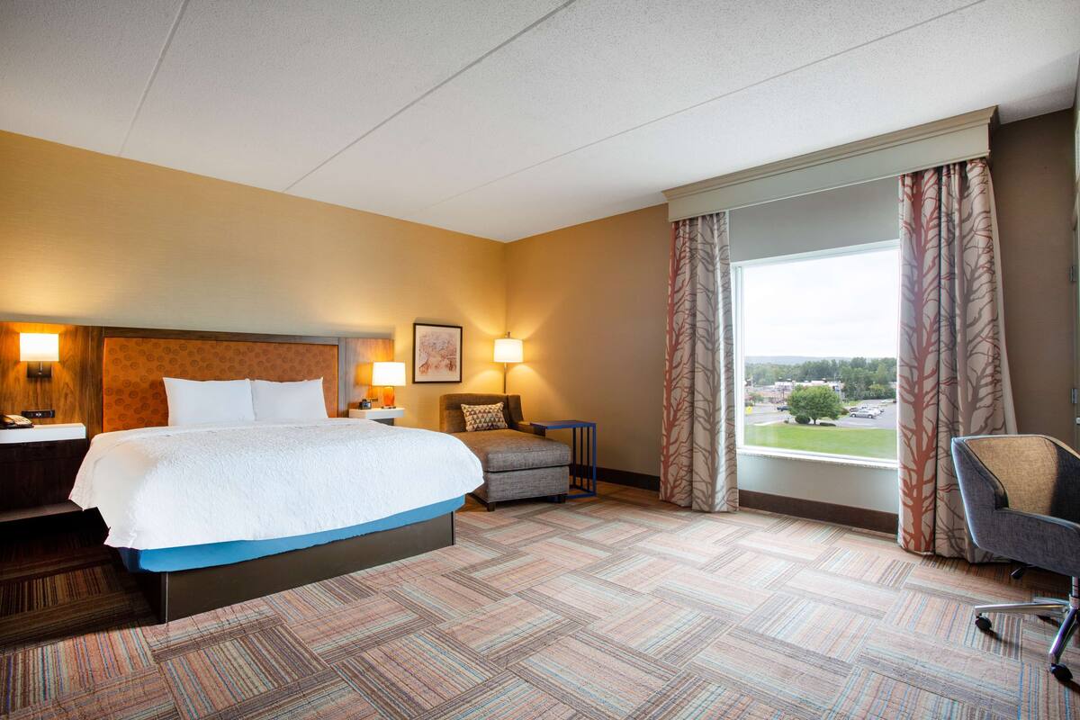 Ein Hotelzimmer mit einem großen Bett, einem Schreibtisch mit Lampe, einem Sessel und Blick ins Freie.