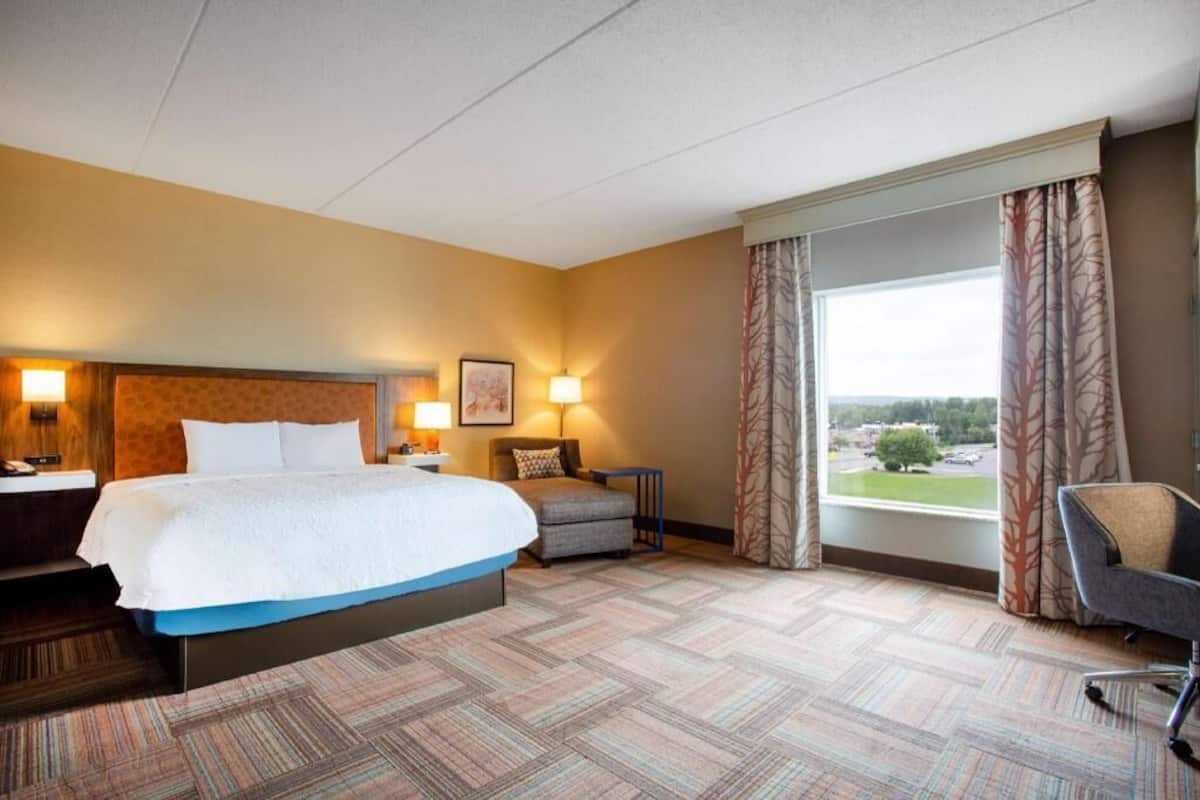 Ein Hotelzimmer mit einem großen Bett, einem Schreibtisch, einem Sessel, einem Fenster mit Vorhängen und Blick nach draußen.