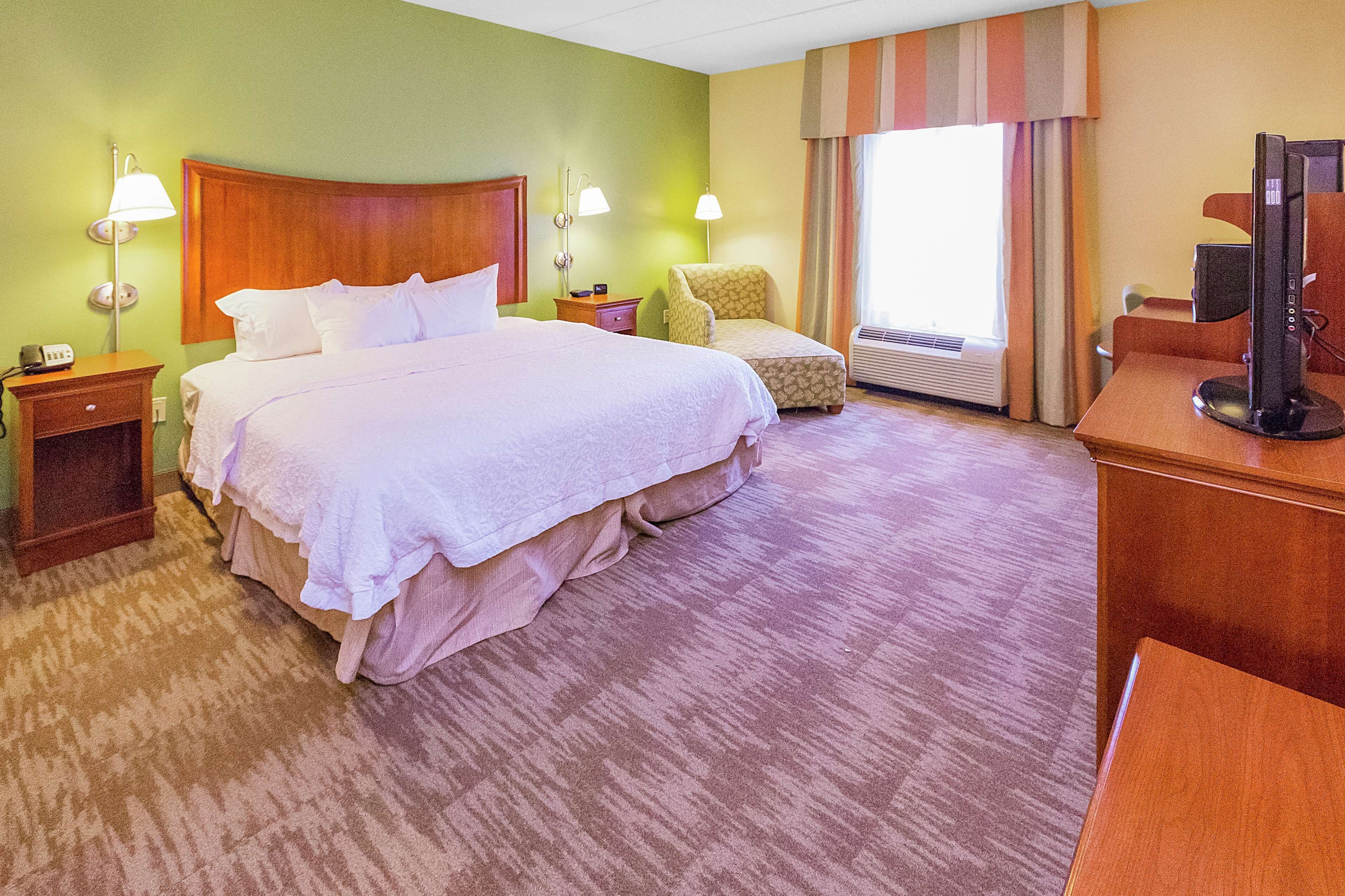 Quarto de hotel com uma cama grande, uma escrivaninha com computador, uma cadeira e uma janela com cortinas.