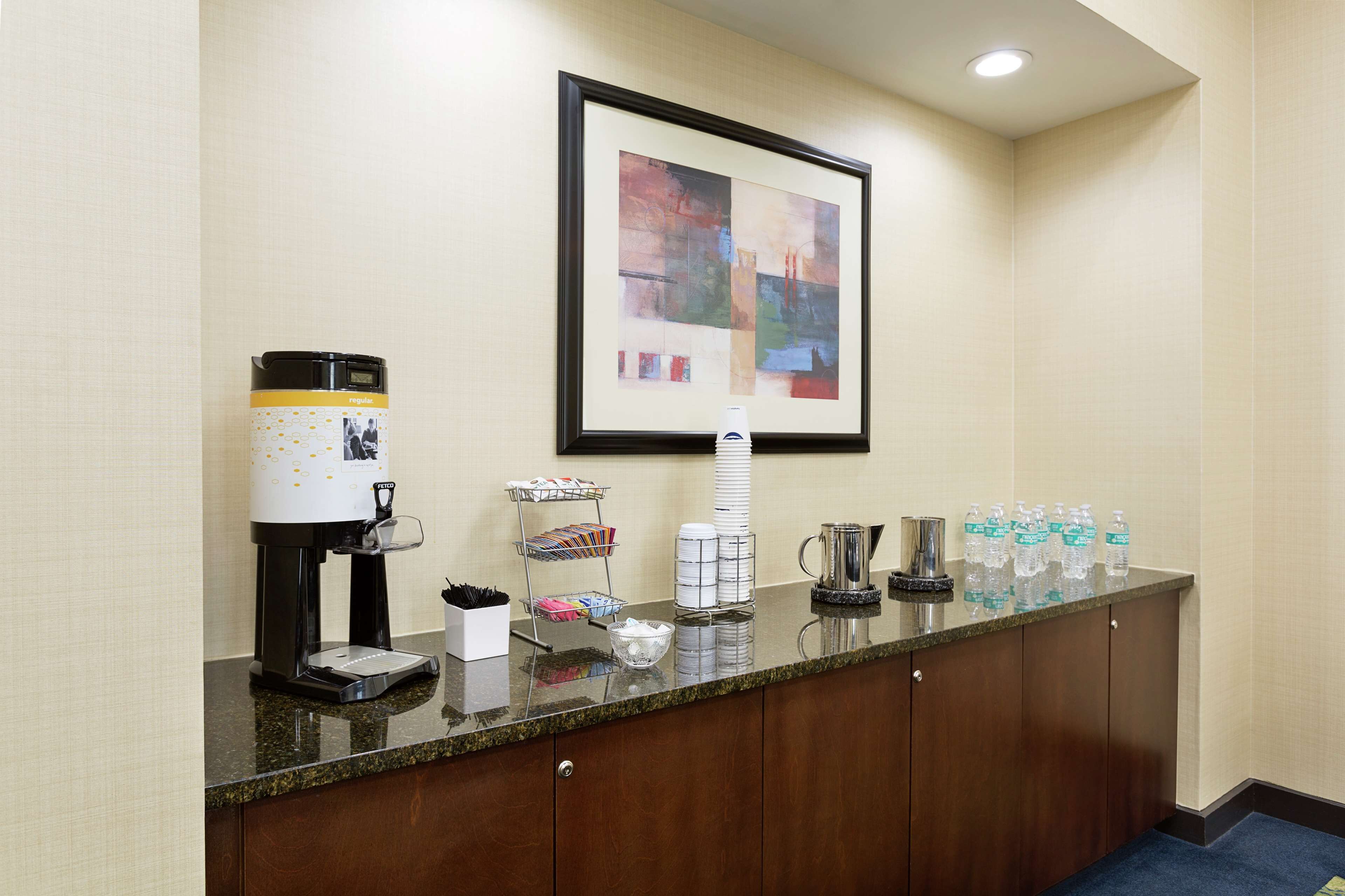 Zona de recepción de hotel con una máquina de café, dispensador de agua y una obra de arte enmarcada en la pared.