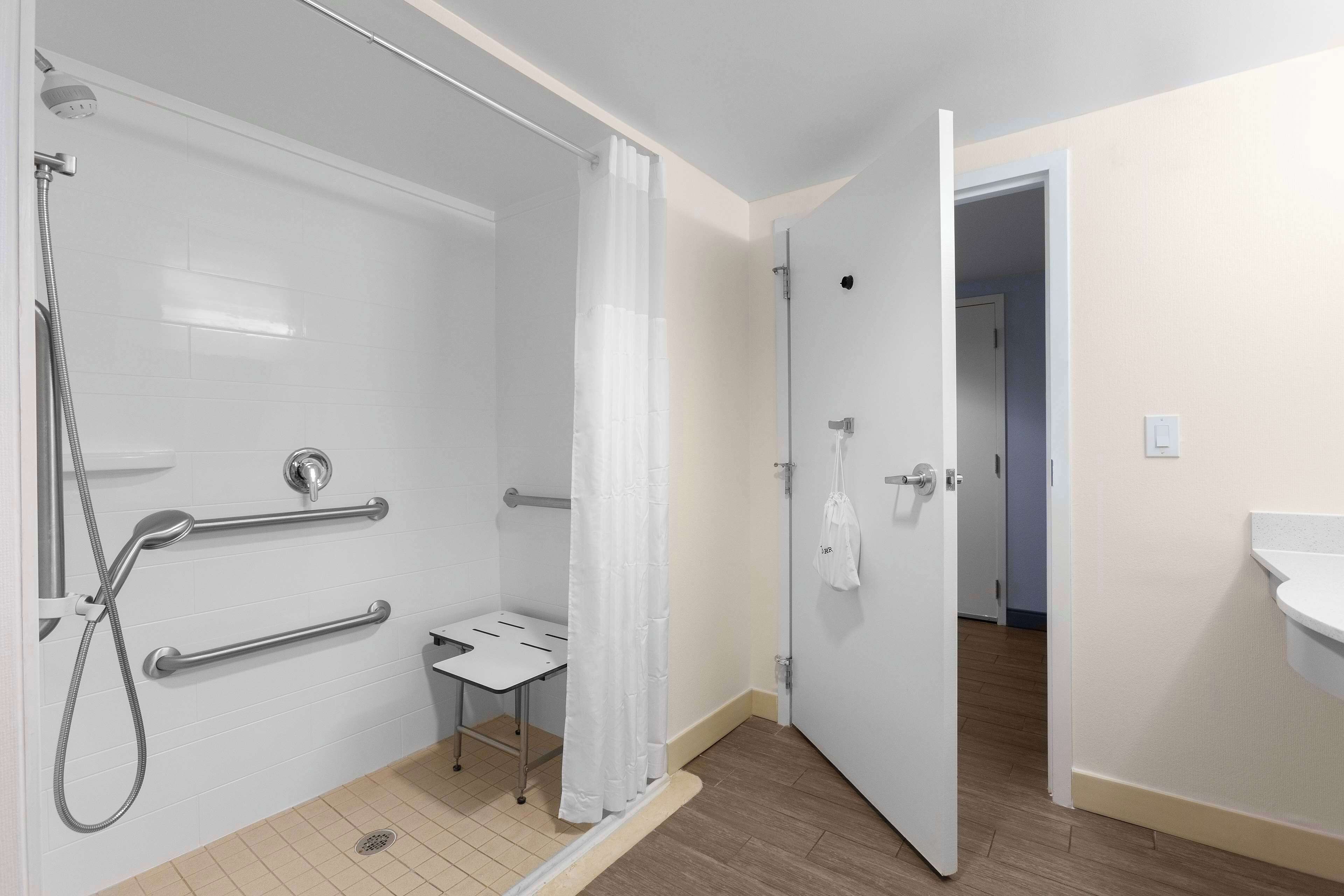 Une salle de bain avec une douche, des barres d’appui, un lavabo blanc et une porte donnant sur une autre pièce.