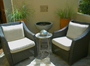 Two wicker chairs with white cushions, a small table with a decorative item, and a large planter with tall plants. - Hotel Cheval (Paso Robles)