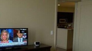 A hotel room with a bed, a dresser, and a television displaying a news broadcast. - Pointe 400 by ExecuStay (St. Louis)