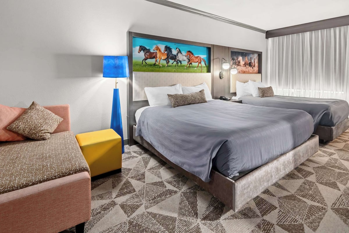 Quarto de hotel com duas camas, um sofá, um abajur e um quadro de cavalos.