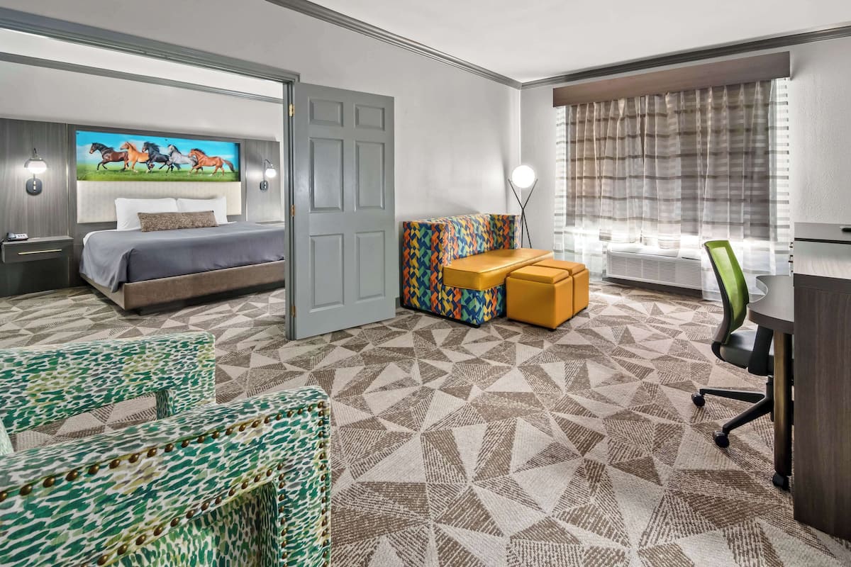 Quarto de hotel com cama, escrivaninha, cadeira, um banquinho colorido e um quadro com cavalos na parede.