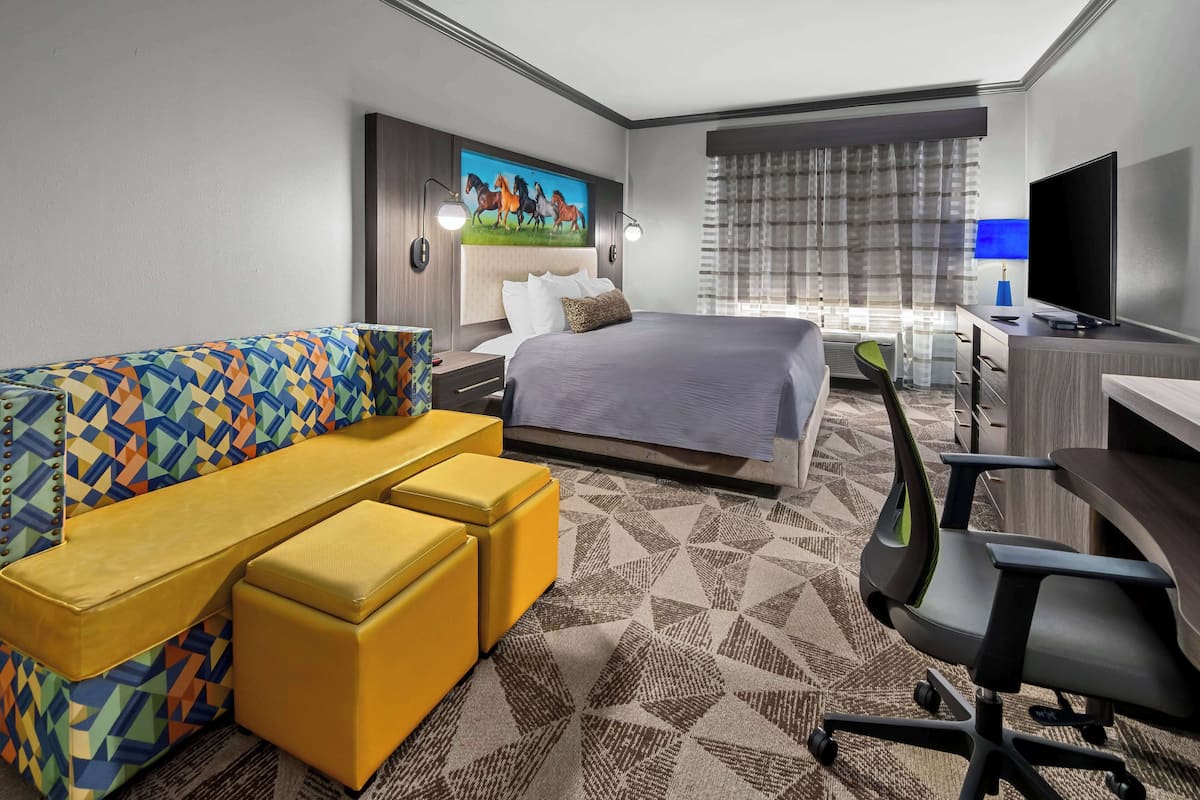 Quarto de hotel com cama, sofá colorido, escrivaninha com cadeira e televisão.