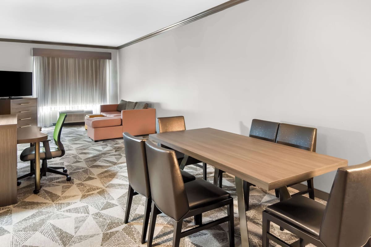 Quarto de hotel com área de refeições, sofá, televisão e janela com cortinas.