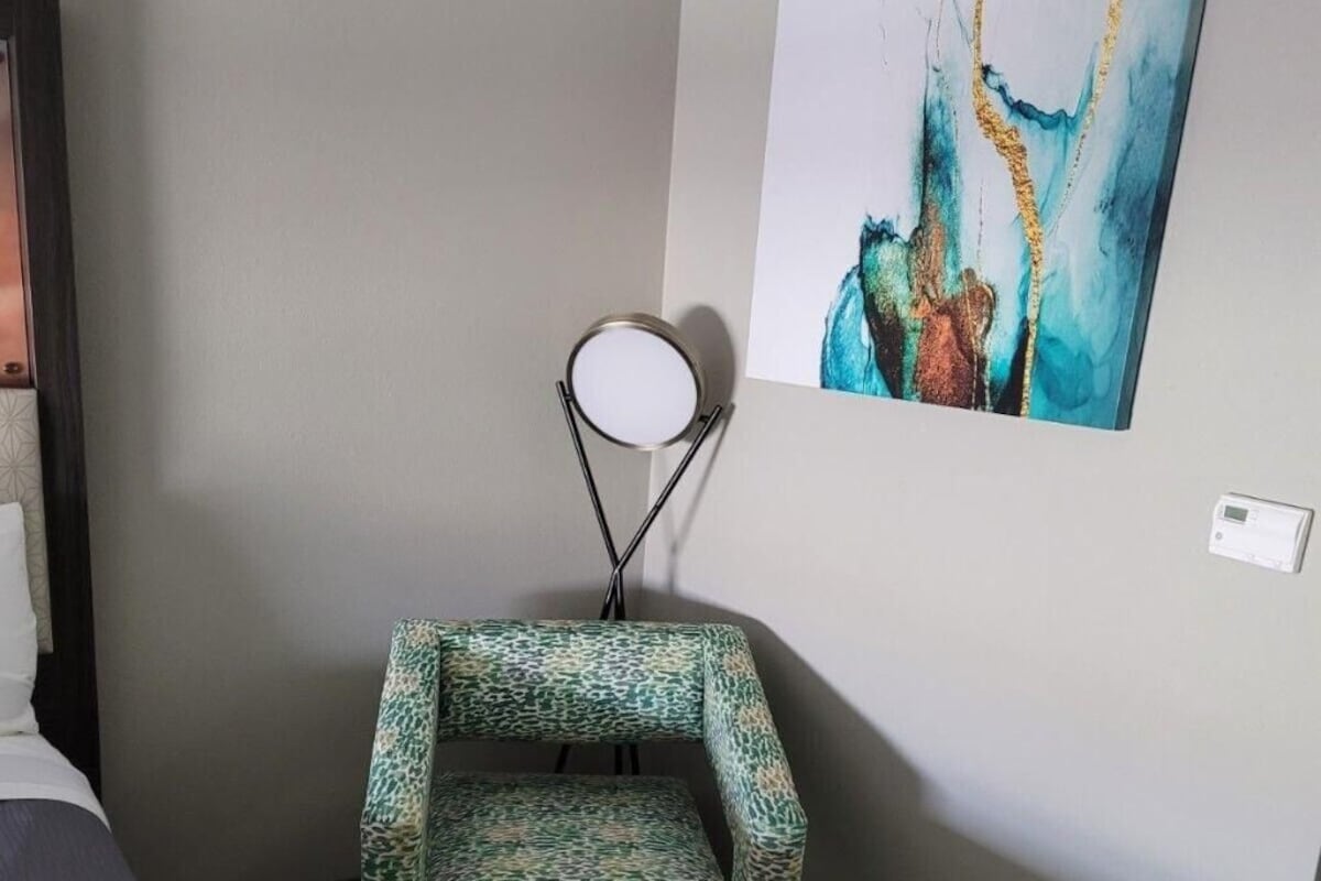 A hotel room with a bed, a patterned armchair, a small ottoman, a wall-mounted lamp, and a painting.